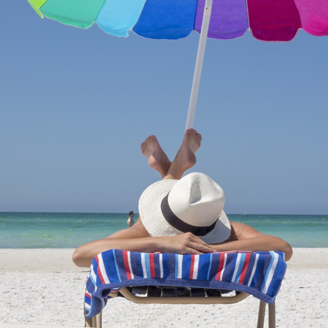 Design ohne Titel Mensch liegt auf einer Liege am Strand unter einem bunten Sonnenschirm und hat einen weißen Sonnenhut auf.